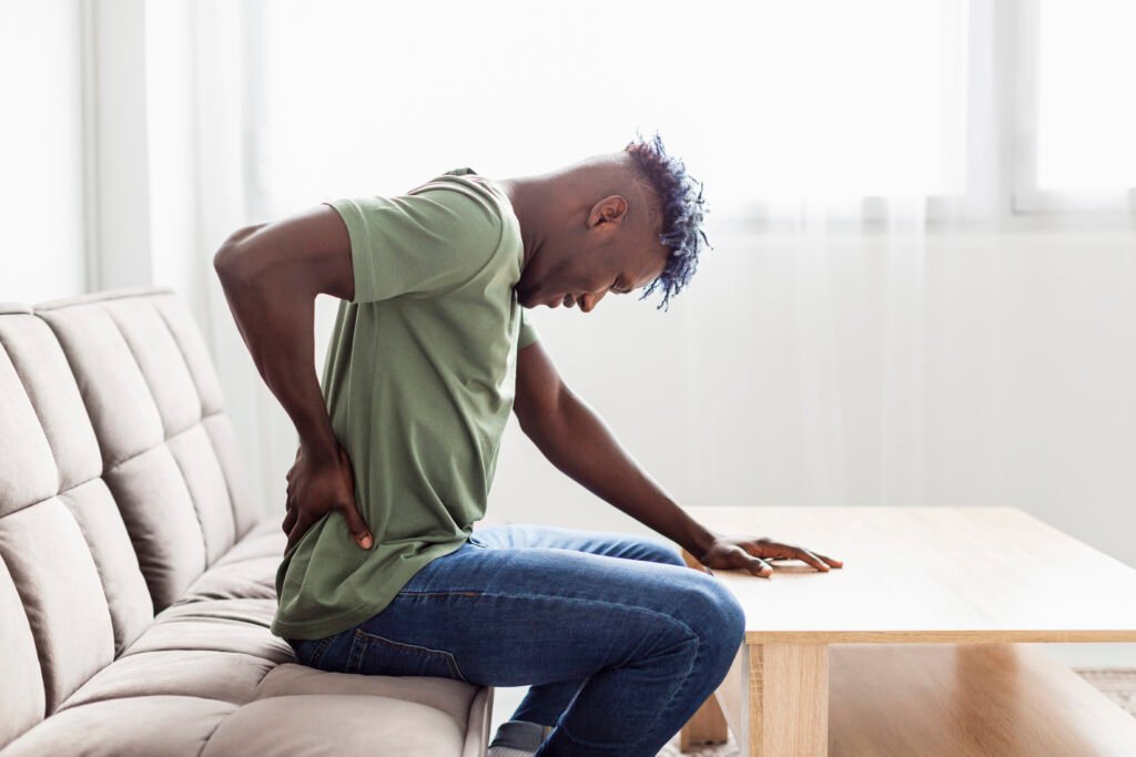 Homem negro, sentado em um sofá, apoiado em uma mesa de centro com as mãos nas costas, demonstrando que está com dores nas costas