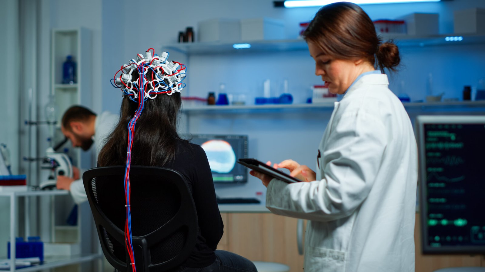 Neurologista com tablet na mão anotando as movimentações cerebrais da paciente, que está sentada na cadeira com uma espécie de touca.