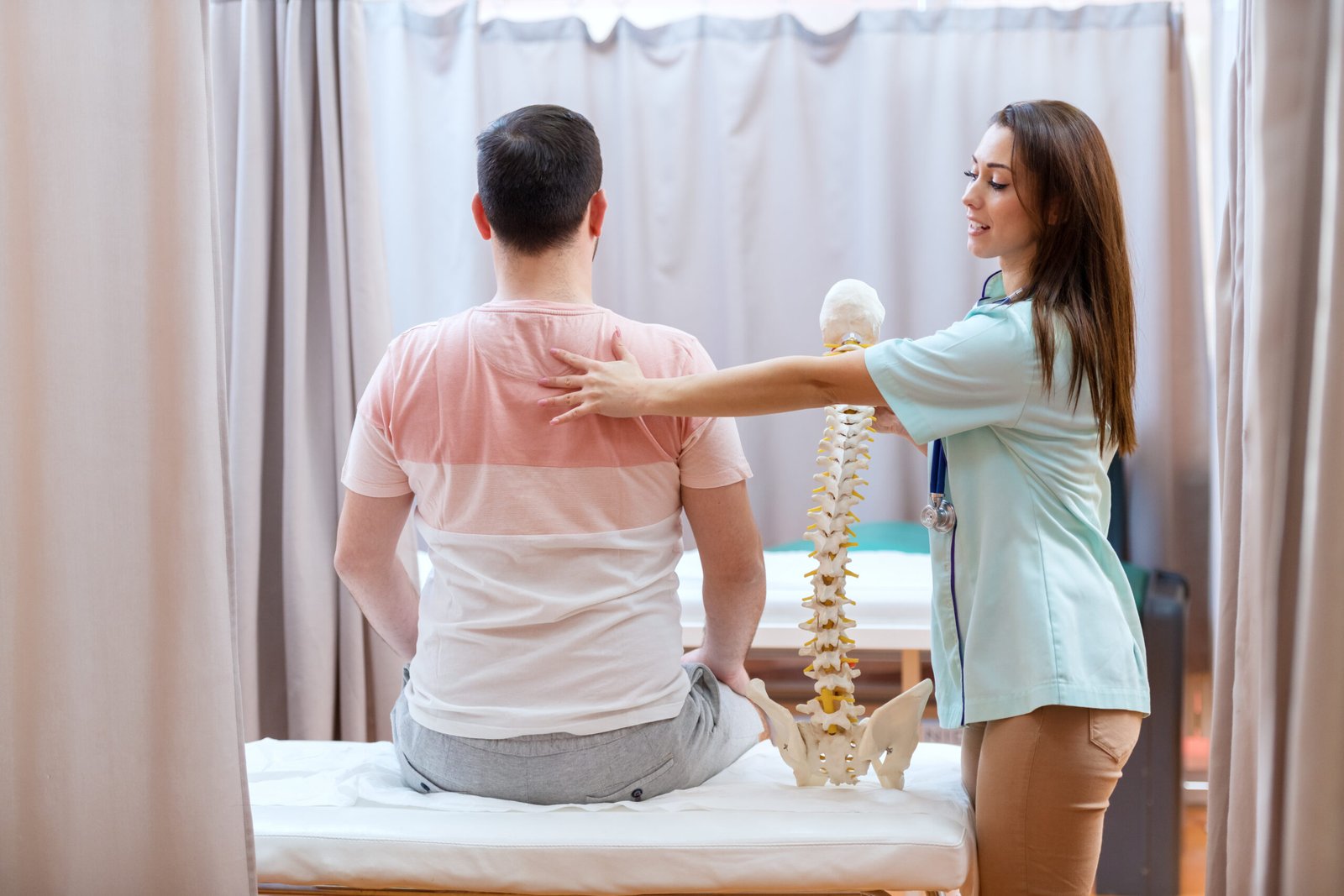 Fotografia de homem sentado em cama no hospital devido a cervicalgia. A enfermeira segura uma estrutura óssea e ao mesmo tempo com as mãos nas costas do paciente.