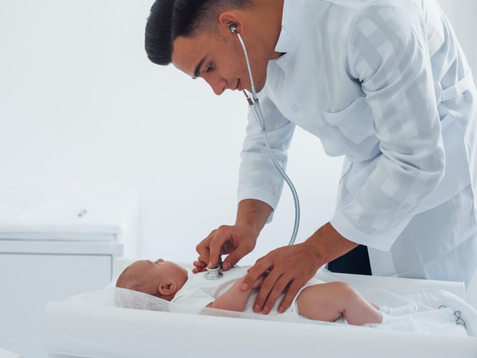 Fotografia de médico realizando exame em um neném. Ele usa o estetoscópio.