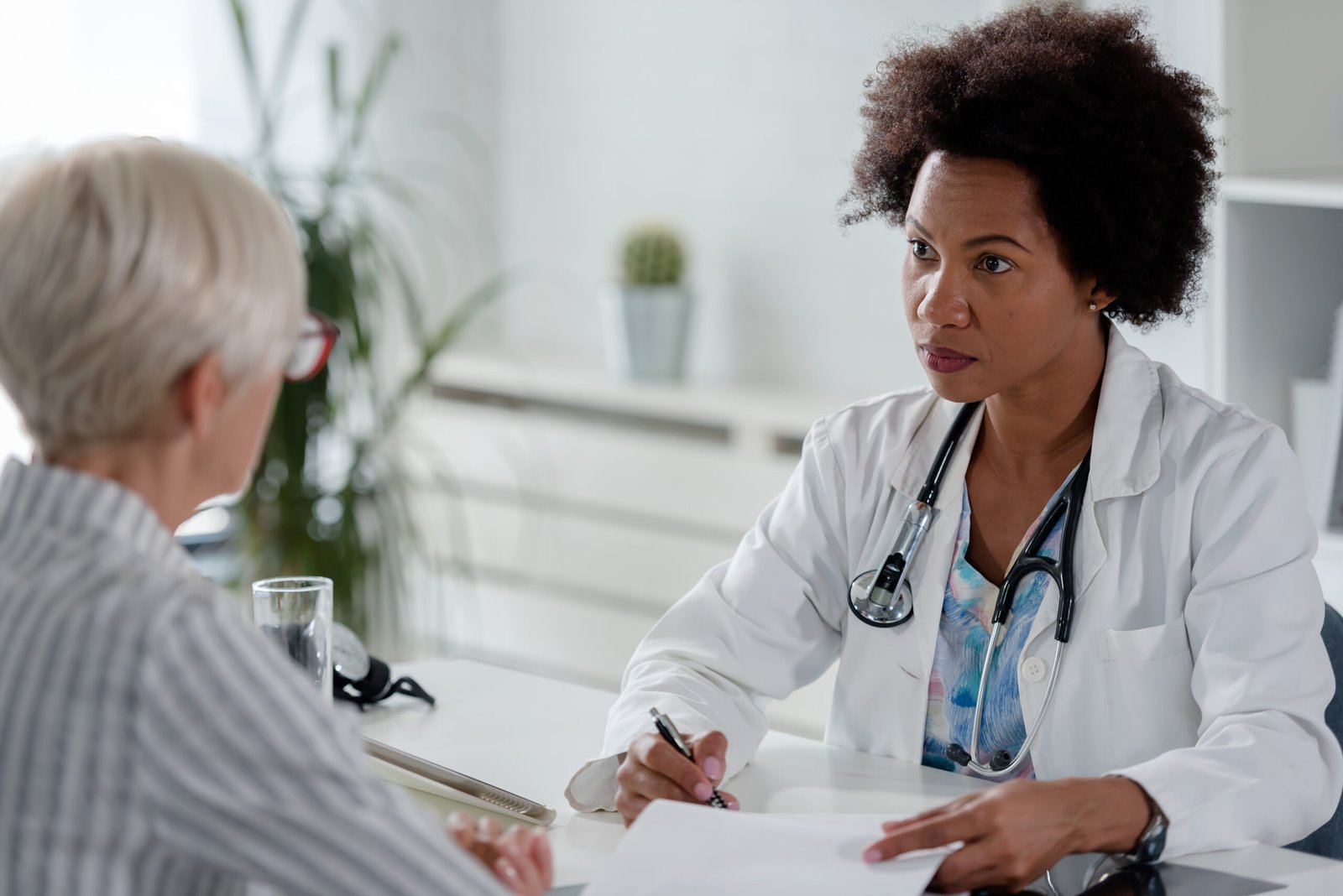 Médica em sua sala examinando paciente com hérnia de disco. A paciente já é idosa, a médica está olhando atentamente para a paciente enquanto está com algumas anotações na mesa.