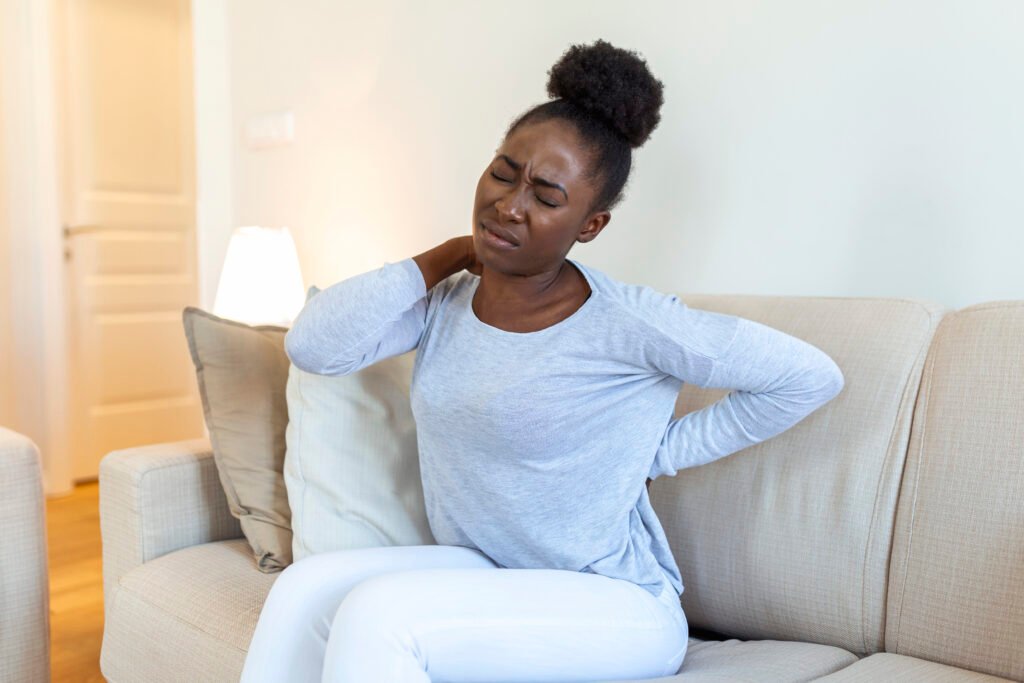 Fotografia de mulher preta sentada num sofá. Ela está com dor neuropática, iniciando no pescoço até o cocix, locais onde está com a mão pressionando. 