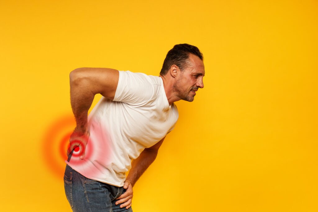 Homem curvada, ainda que pouco, com a mão direita na lombar. Ele está com expressão de dor, há uma grande probabilidade de estar com a dor gerada da lombar alterada.