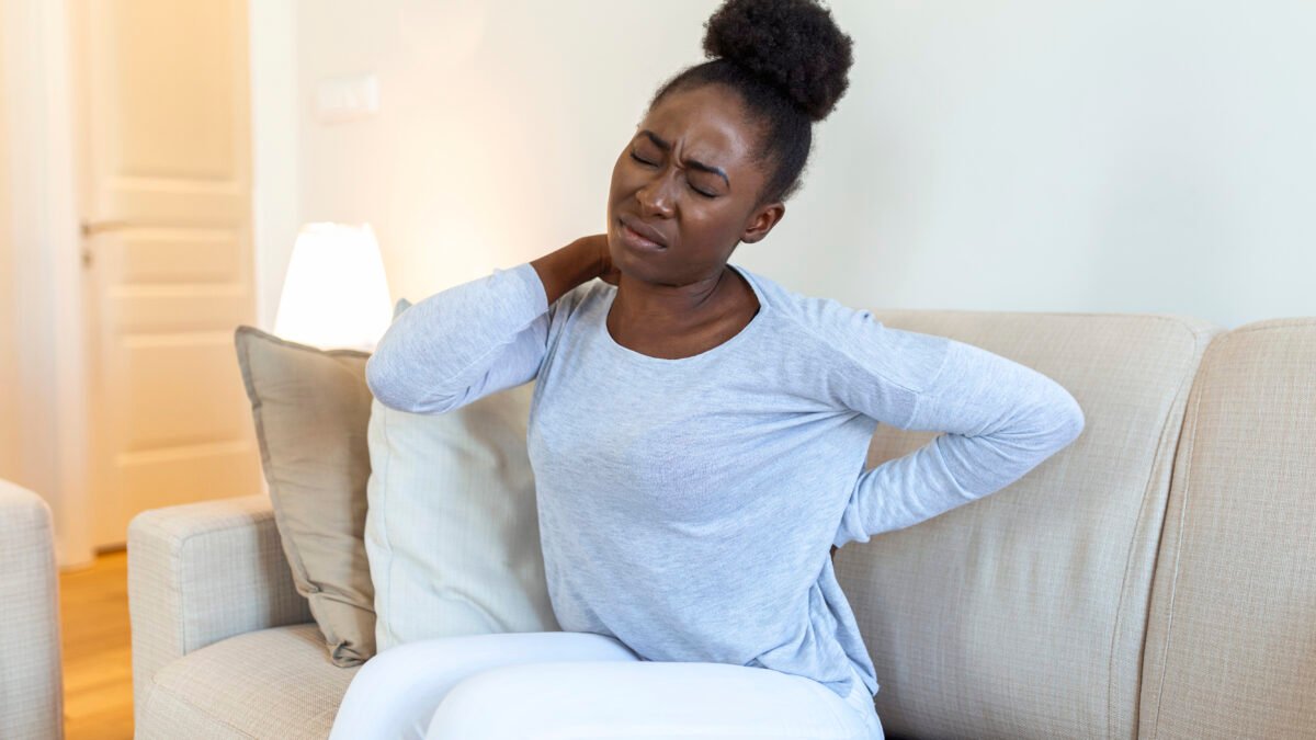 fotografia de mulher negra com expressão de dor. Ela está com dor no pescoço e nas costas, pois está tentando massagear com as mãos. Ela está sentada no sofá de sua casa.