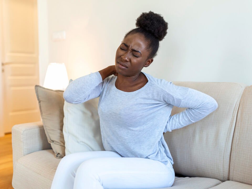 fotografia de mulher negra com expressão de dor. Ela está com dor no pescoço e nas costas, pois está tentando massagear com as mãos. Ela está sentada no sofá de sua casa.