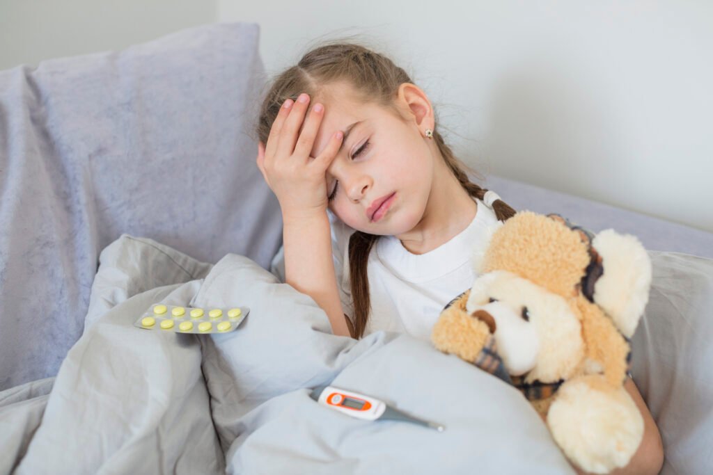 Fotografia de uma menina de 7 anos sentada na cama. Ela está com os olhos fechados e com a mão na cabeça, indicando estar com dor. Também há remédio sob a coberta.