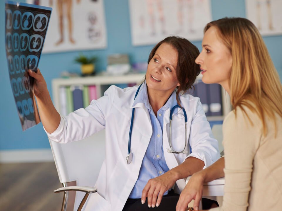 Fotografia de médica mostrando exames para a paciente, que buscou um profissional para descobrir se possui esclerose múltipla. Elas estão numa sala de consultório médico privado.