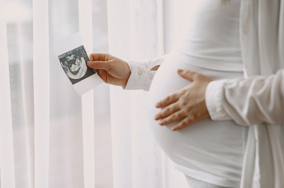 Fotografia de mulher grávida com a mão na barriga e na outra mão segura uma fotografia do ultrassom