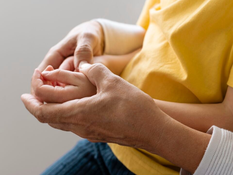 Fotografia de mão de adulto segunda a mão se uma criança com a Síndrome de Apert. A criança apresenta o sintoma Sindactilismo, que são os ósseos dos dedos unidos.