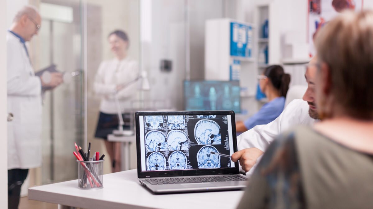 Fotografia de mulher sendo atendida por um neurologista. Na imagem, mostra um notebook com exames de imagem do cérebro do paciente.