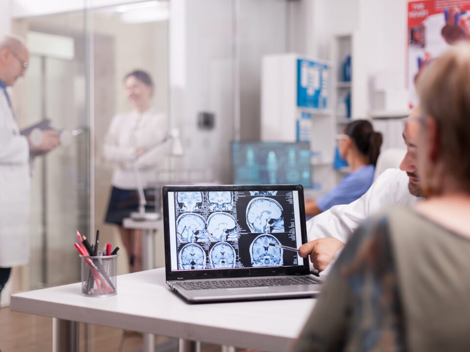 Fotografia de mulher sendo atendida por um neurologista. Na imagem, mostra um notebook com exames de imagem do cérebro do paciente.