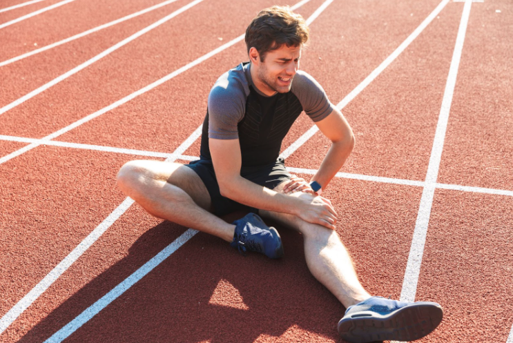 Fotografia de homem sentado no chão de um ambiente para corrida. Ele está com expressão de dor enquanto toca o joelho direito.