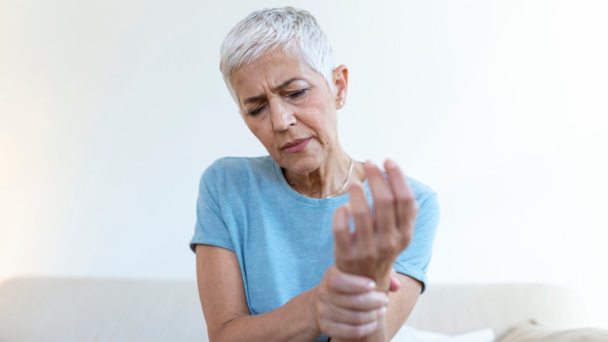 Fotografia de uma mulher com mais de 60 anos. Ela está com uma expressão de incômodo, enquanto olha e segura o braço direito, mais próximo do pulso.