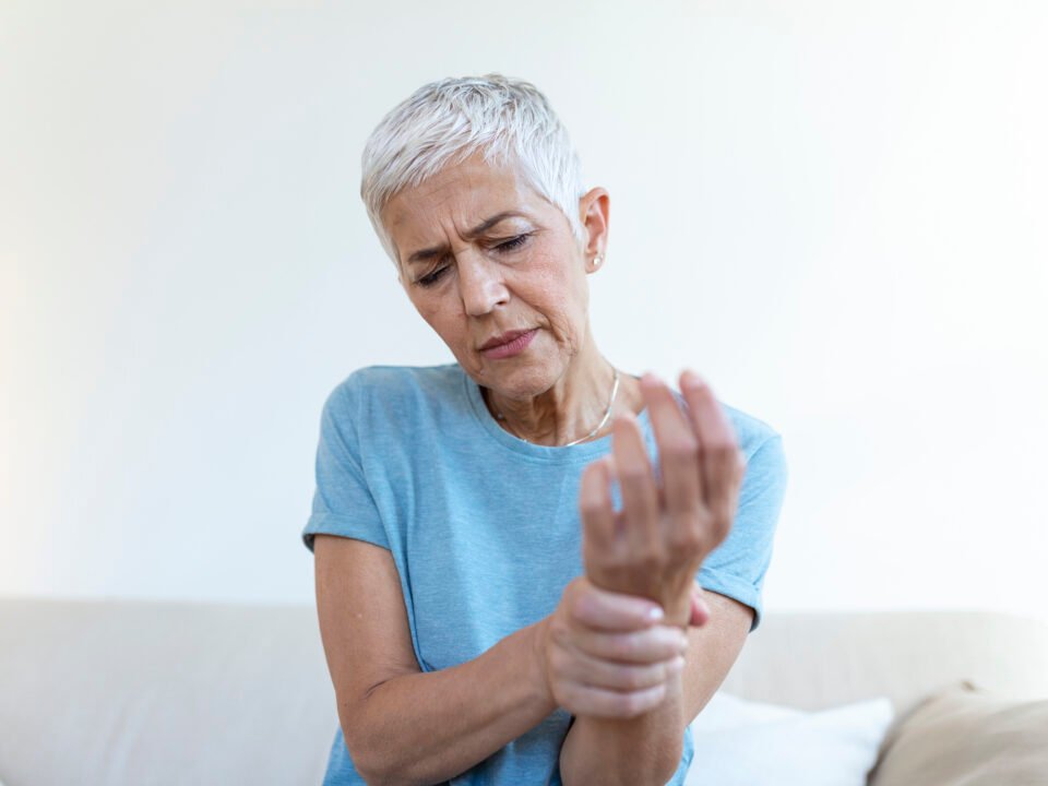 Fotografia de uma mulher com mais de 60 anos. Ela está com uma expressão de incômodo, enquanto olha e segura o braço direito, mais próximo do pulso.