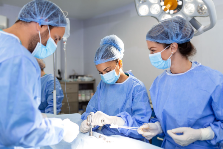 Fotografia de cirurgiões realizando um procedimento. Na iamgem mostra três médicos, duas mulheres e um homem na sala de cirurgia.