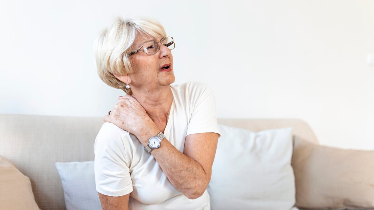 Fotografia de mulher em torno de 60 anos sentada no sofá. Ela está com a mão no ombro, com expressão de dor.