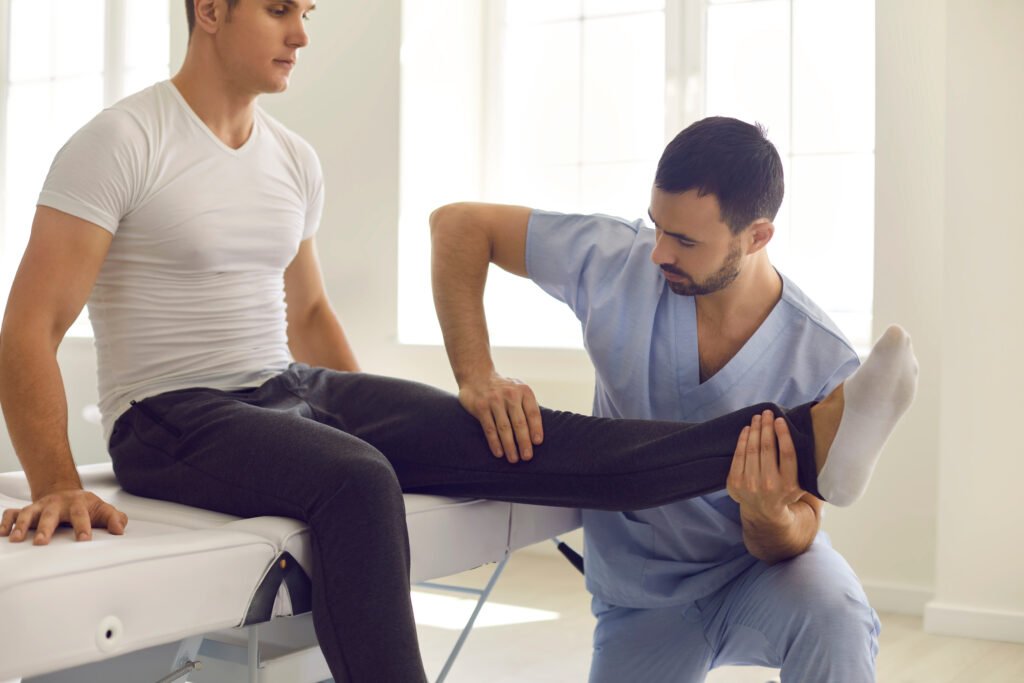 Fotografia de profissional da saúde auxiliando um paciente masculino. Ele está esticando a perna direita do paciente, que está sendo na cama do hospital.