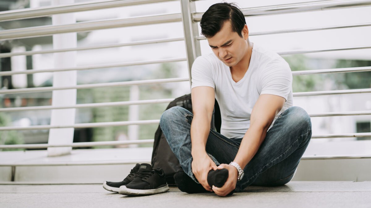 Imagem de homem sentado no chão, sem os sapotos nos pés. Ele está tendo uma cãibra no pé direito, sua expressão é de dor enquanto ele pressiona o local.