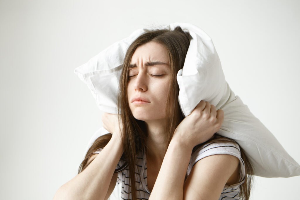 Fotografia de mulher sentada, com o travesseiro no pescoço e expressão de raiva por não estar conseguindo dormir.