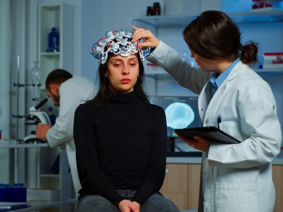 Fotografia de mulher e médica. A médica neurologista está fazendo exames cerebrais na paciente, utilizando instrumentos como um capacete de fios, para mapeamento cerebral.