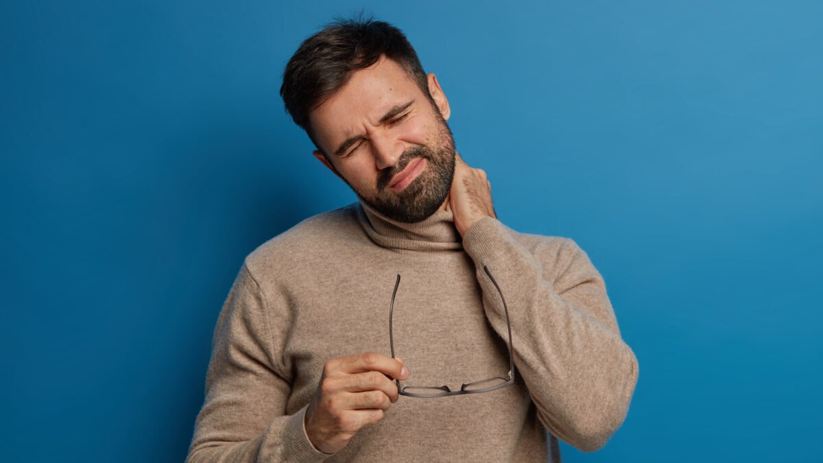 Fotografia de homem com dor no pescoço, podendo ser um sintoma da espondilose cervical. Ele está com uma mão na nuca e a outra segurando o óculos, sua expressão é de dor.