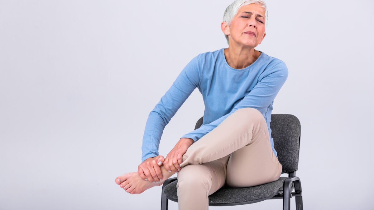 Fotografia de mulher, aproximadamente 60 anos, sentada numa cadeira enquanto faz uma compressão com as mãos no próprio pé. Sua expressão é de dor ou incômodo.