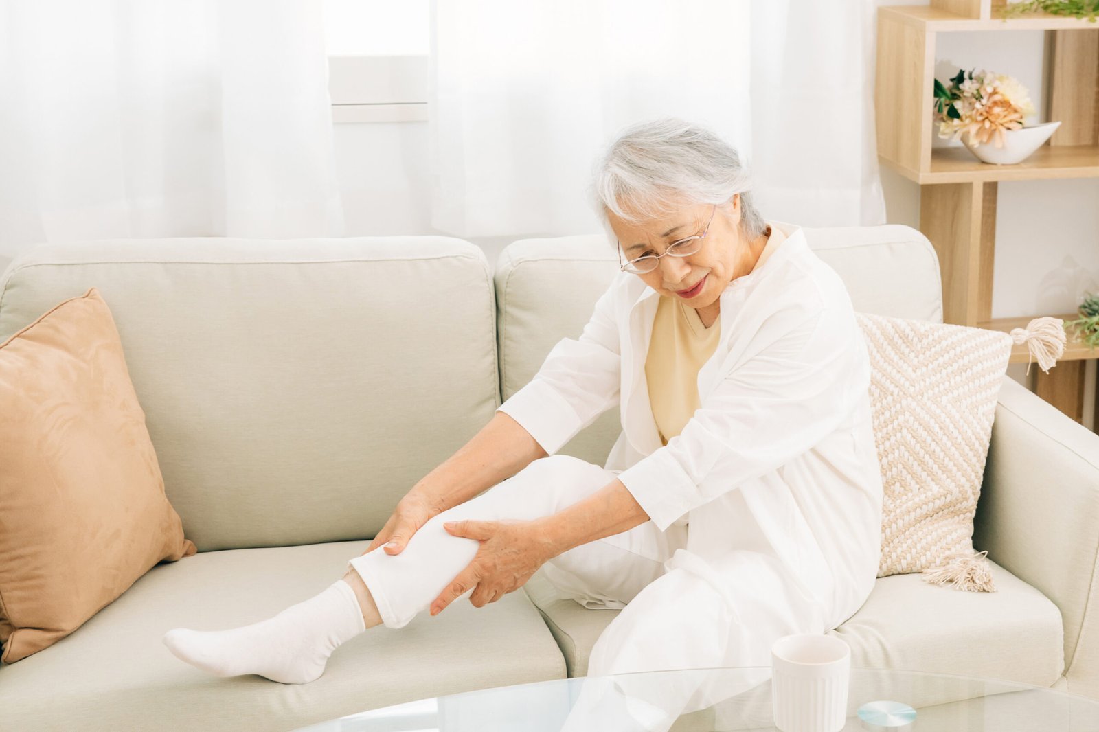 Fotografia de idosa sentada em seu sofá. Ela está com a perna direita sobre o sofá enquanto passa a mão na região na panturrilha. Sua expressão é de dor.