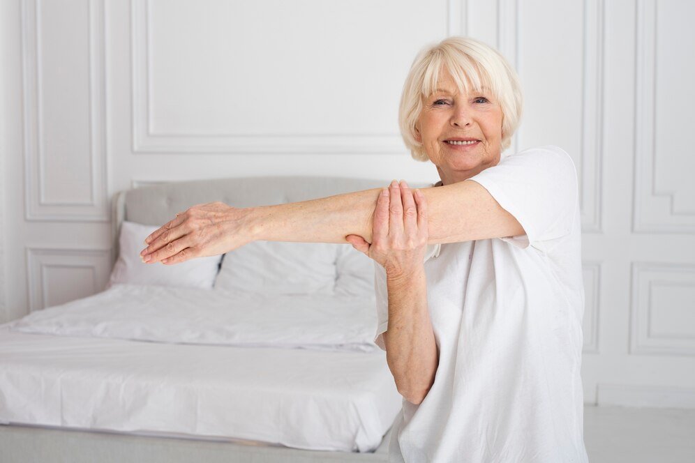 Foto de uma mulher idosa alongando seu braço direito na recuperação pós artrodese cervical. Ela veste roupas brancas e está em um quarto branco.